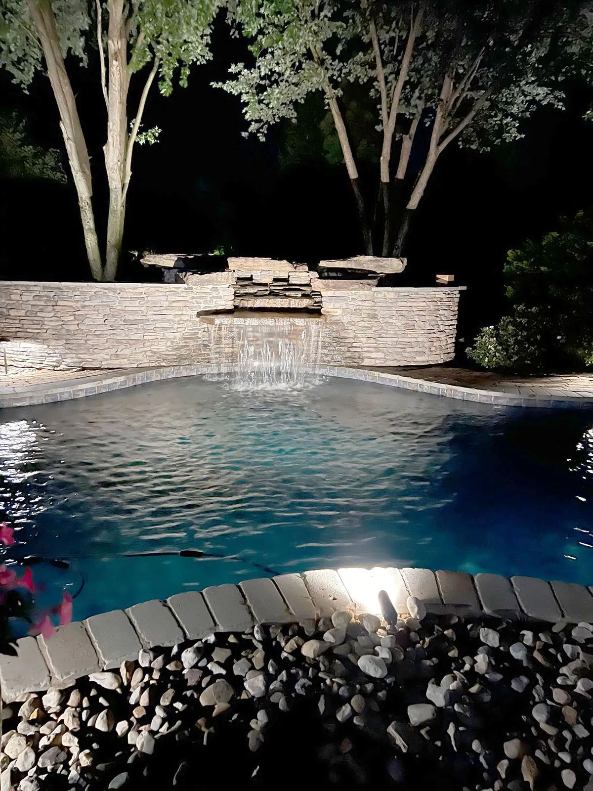 A well-lighted swimming pool with a waterfall in the backyard at night
